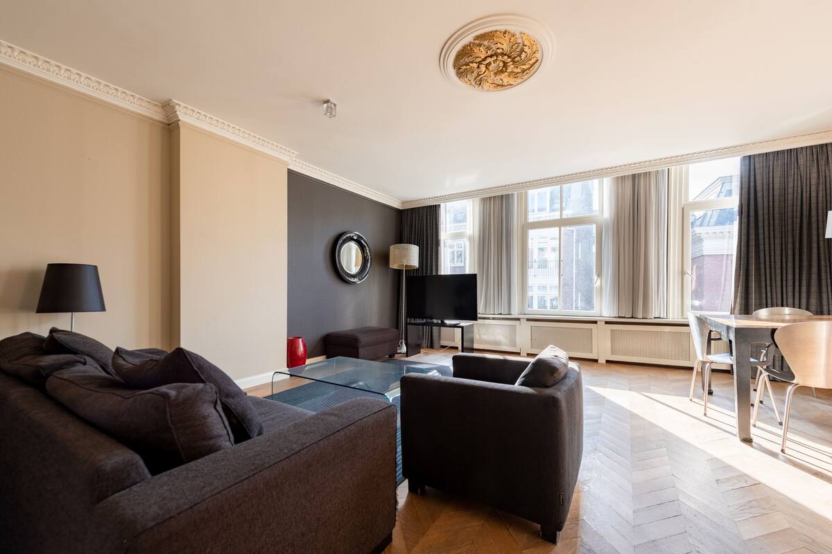 A modern living room with a sofa, armchair, and a glass coffee table. There is a flat-screen TV, a lamp, and a dining table with chairs.
