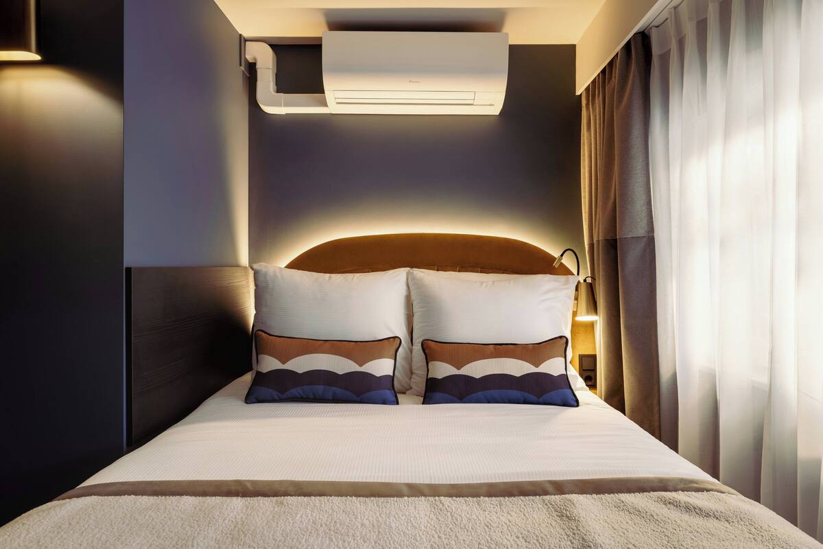 A hotel room with a bed, pillows, an air conditioning unit, and curtains.