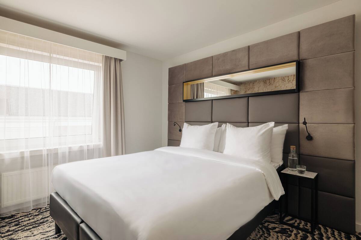 A hotel room with a large bed, a bedside table with a lamp, a window with sheer curtains, and a headboard with a mirror.