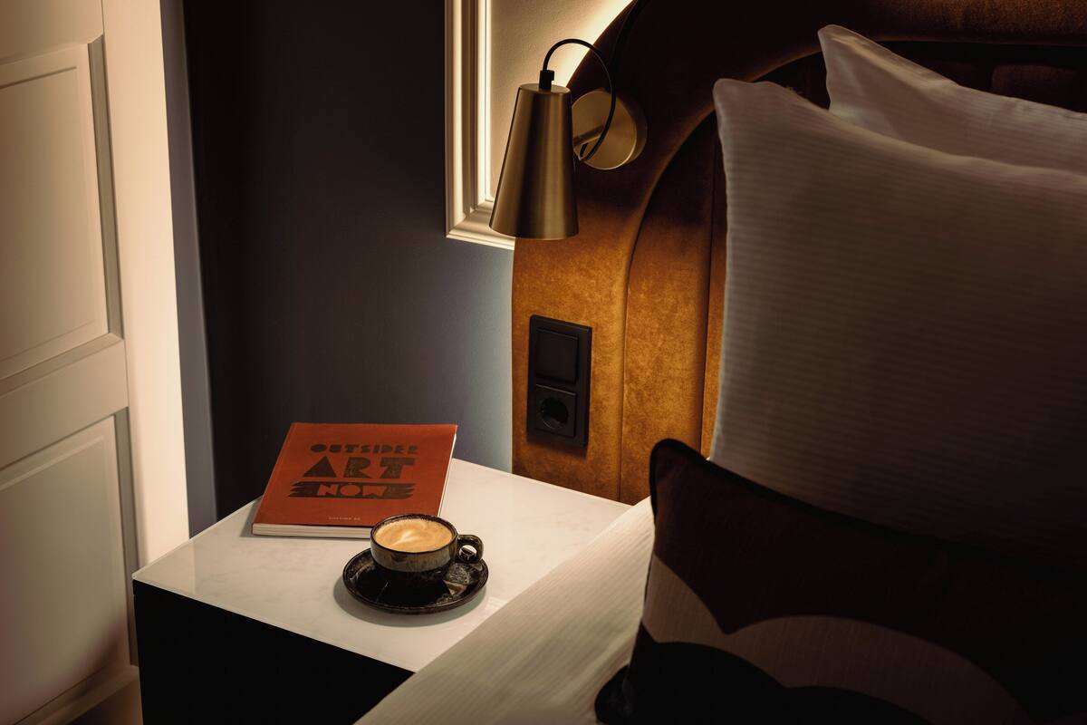 A hotel room with a bed, a bedside table with a book and a cup of coffee, a lamp, and a window.