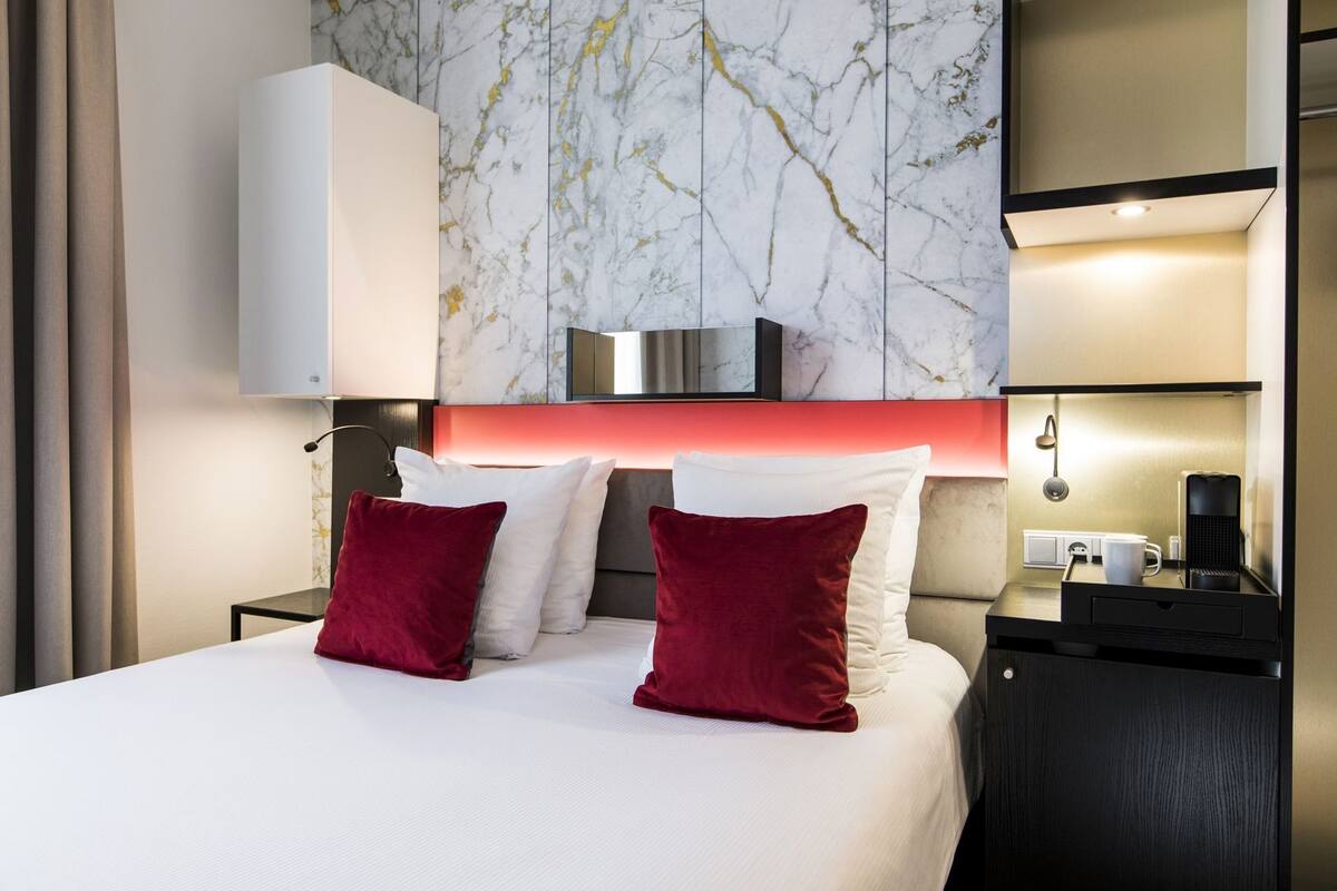 A hotel room with a bed, red pillows, a nightstand, a coffee maker, and a marble wall.