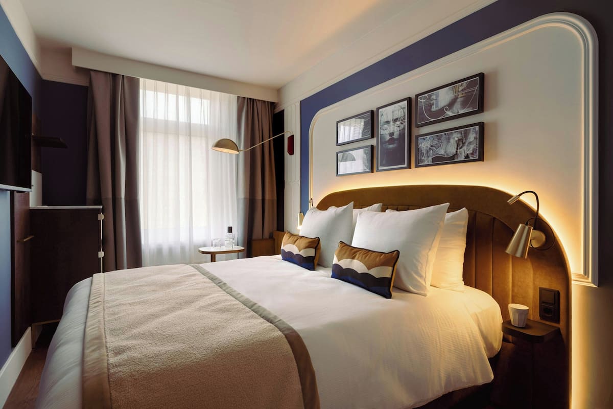 A hotel room with a large bed, a bedside table, a lamp, and a window with curtains.