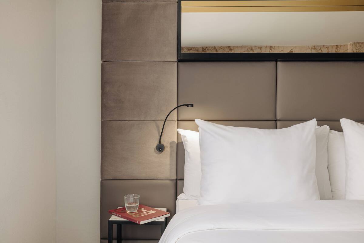 A neatly made bed with white linens, a headboard with a light brown panel, and a small bedside table with a book and glass of water.