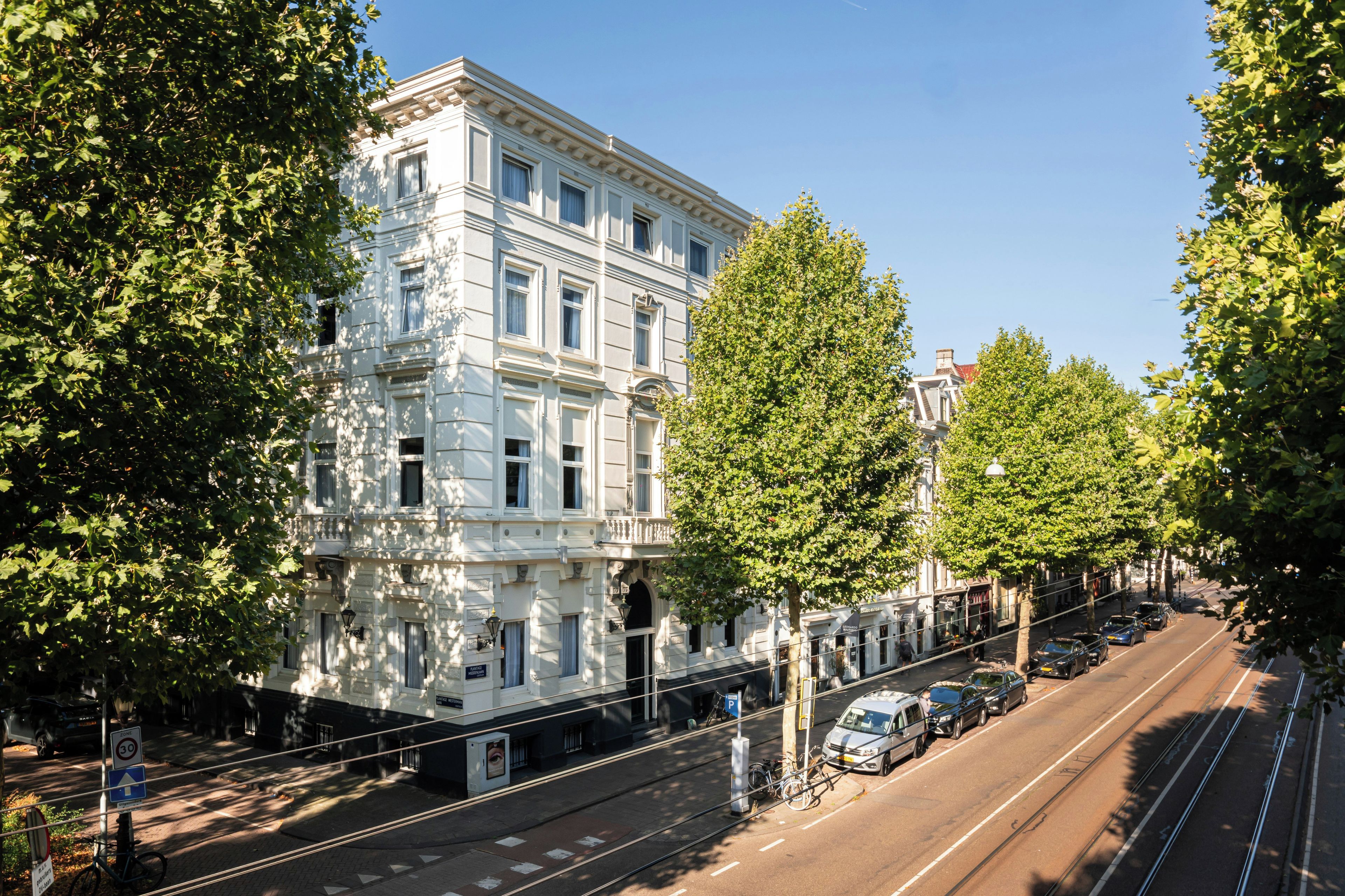 A street view with a row of parked cars, a tram line, and a building with classical architecture.