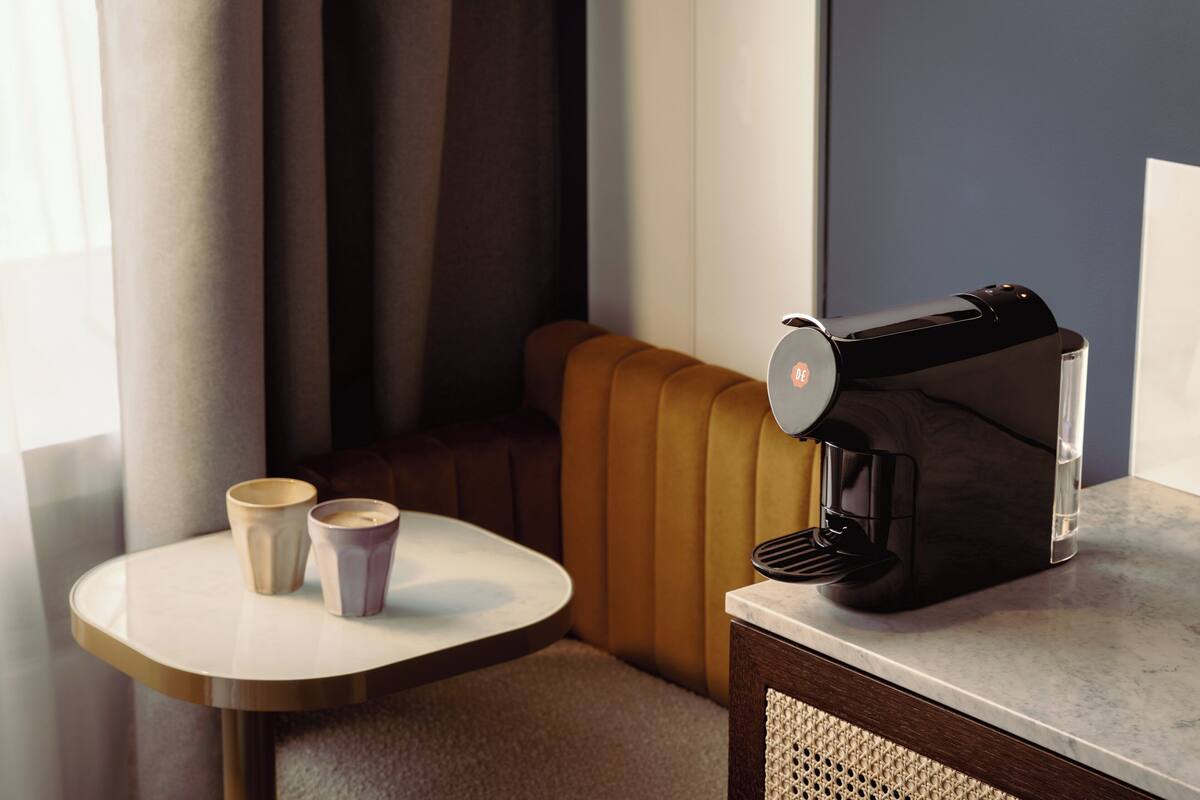 A coffee machine on a table with two cups, a cushioned bench, and a window with curtains.