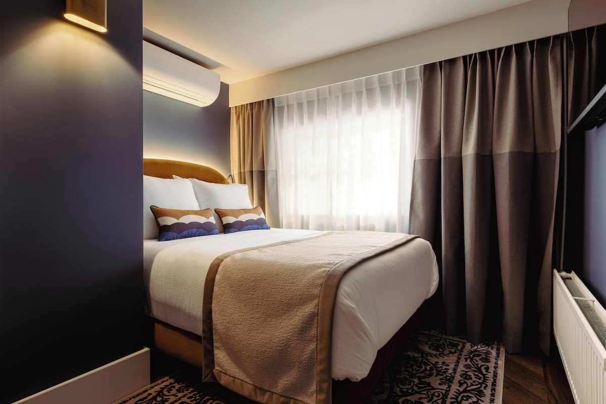 A hotel room with a bed, a window with curtains, a radiator, and a patterned rug.