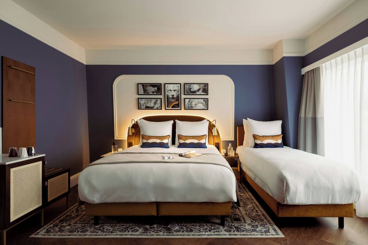 A double bed with a headboard, bedside tables with lamps, a large window with curtains, and artwork on the wall.
