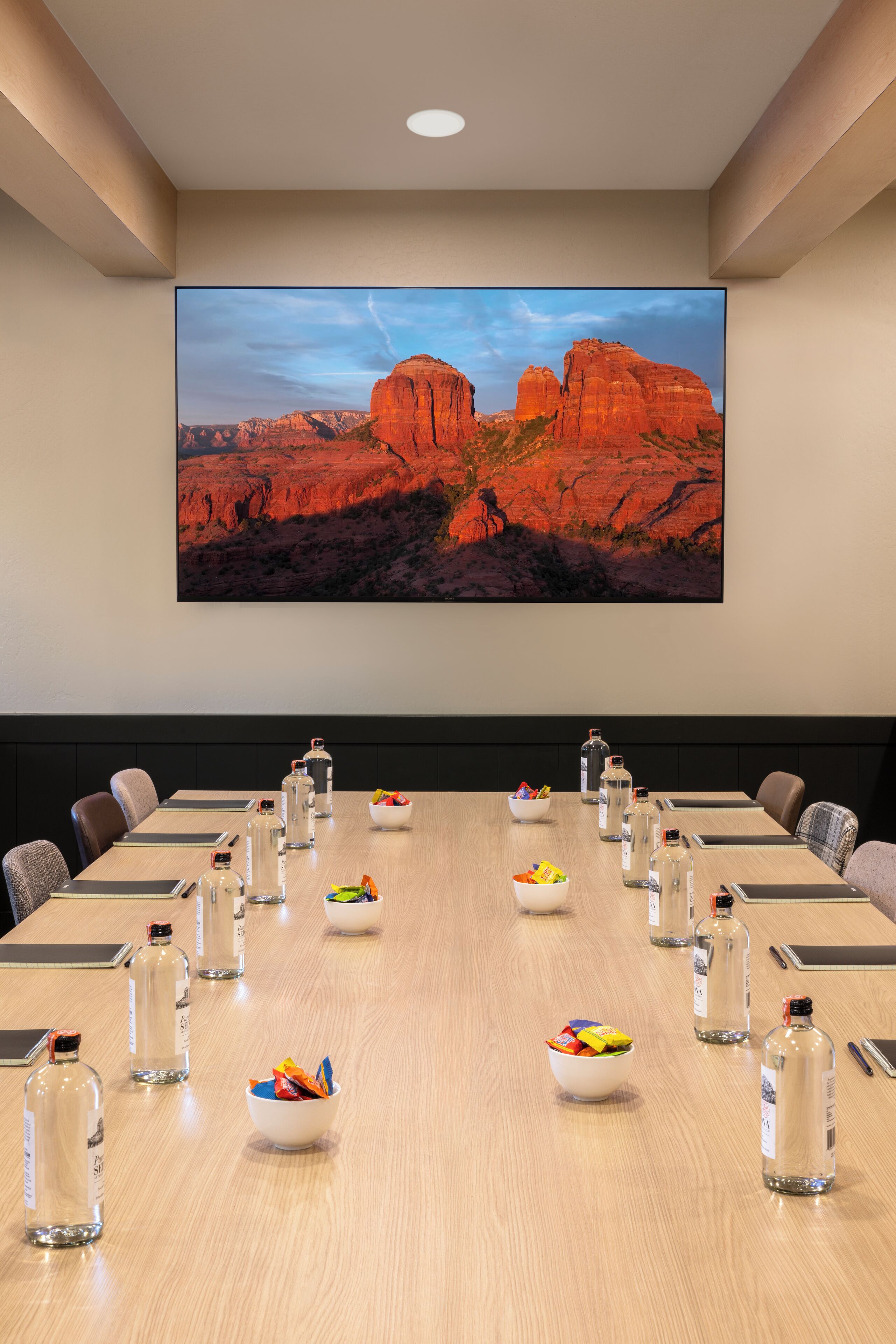 A conference room with a large desert landscape painting, a long table with chairs, and various items like water bottles and snacks.