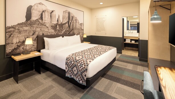 A hotel room with a large bed, a nightstand, a lamp, a wall-mounted TV, and a bathroom visible through an open door. - Mountain Modern Sedona, Tapestry Collection by Hilton (Sedona)