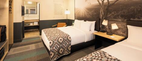 A hotel room with a large bed, a smaller bed, a desk with a lamp, a mirror, and a view of a tree and landscape.