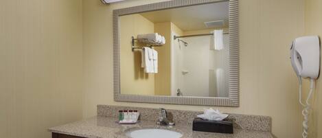A hotel bathroom with a granite countertop sink, a mirror, a towel rack, a hair dryer, and a bottle of hand soap.