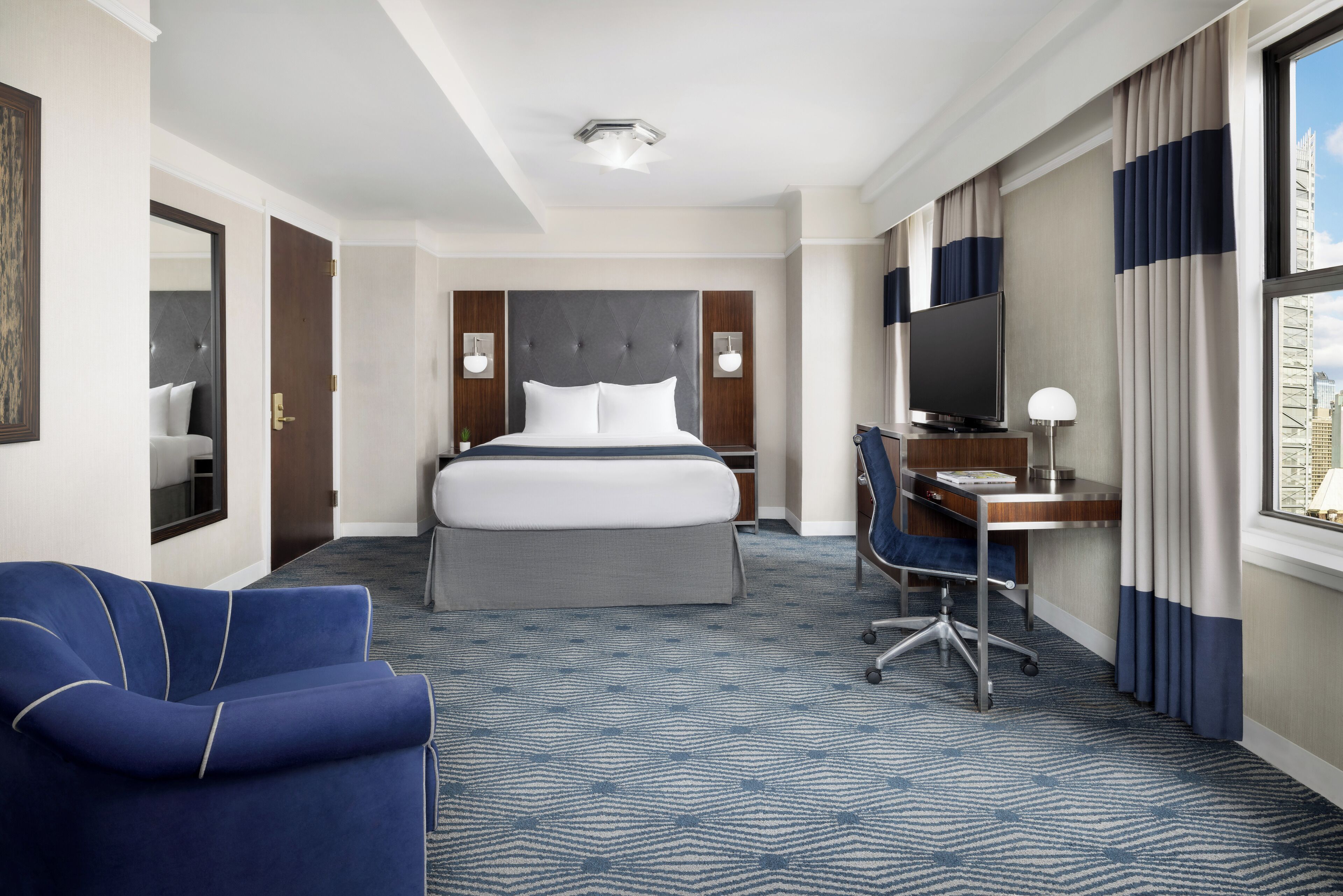 Una habitación de hotel con una cama grande, un escritorio con una computadora, un sillón azul y vistas a la ciudad desde la ventana.