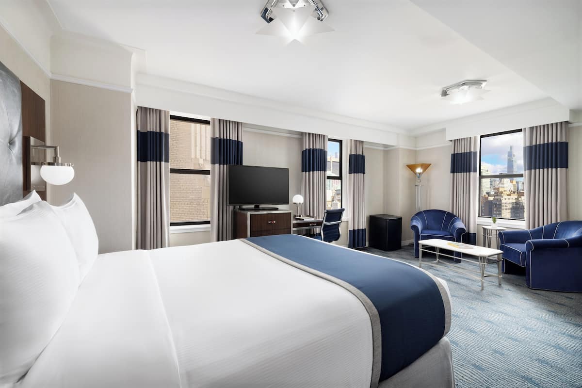 A hotel room with a large bed, a desk, a chair, a TV, and a view of the city.