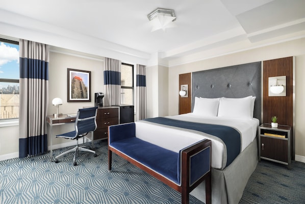 A hotel room with a large bed, a desk with a chair, a small side table, and a window with curtains.