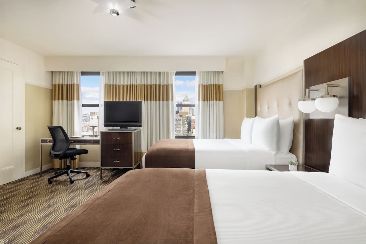 A hotel room with two beds, a desk, a TV, and a city view.