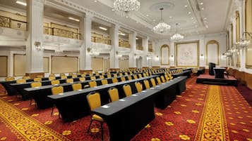 Konferenssihuone, jossa on riveissä pöytiä ja tuoleja, koristeellisia kattokoristeita ja suuria kattokruunuja.