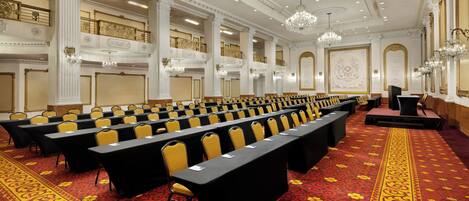 Un salón de conferencias con filas de mesas y sillas, detalles ornamentales en el techo y candelabros.