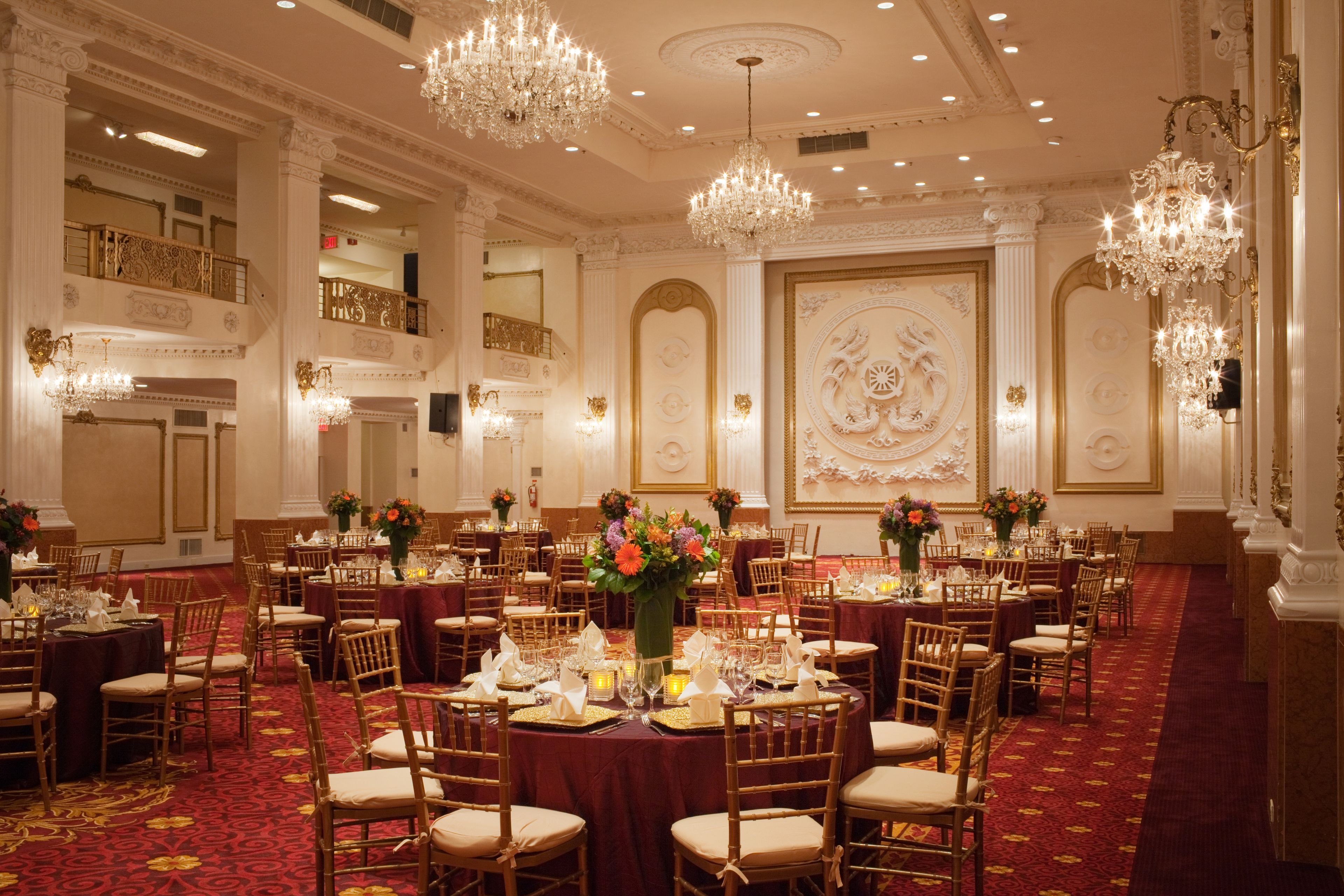 Un salón de banquetes amplio con varias mesas redondas, candelabros ornamentales y un suelo cubierto de alfombra roja.