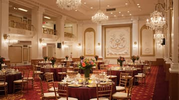 Un salón de banquetes amplio con varias mesas redondas, candelabros ornamentales y un suelo cubierto de alfombra roja.