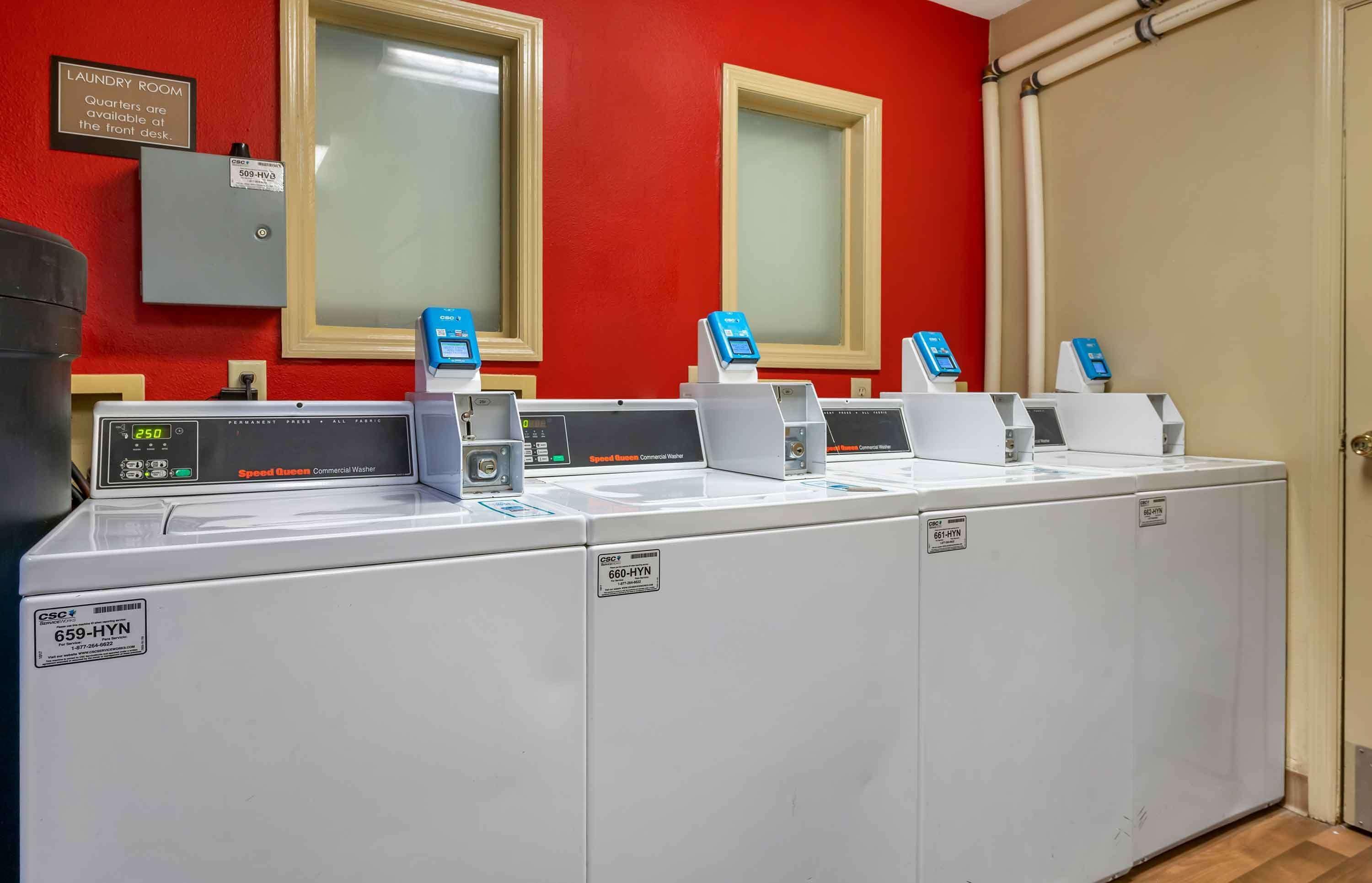 A laundry area with four commercial washers, a speed screen, and a sign indicating quarters are available at the front desk.