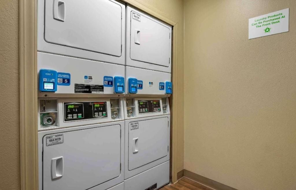 A laundry facility with multiple washing machines and dryers, a sign indicating laundry products can be purchased at the front desk, and a control panel with digital displays.