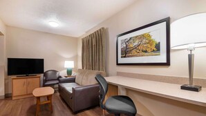 A hotel room with a flat-screen TV, a sofa, a desk with a lamp, a chair, a small table, and a framed picture on the wall. - Studio 6 Corporate Extended Stay Indianapolis, IN - North Carmel (Carmel)
