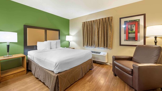 A hotel room with a large bed, two bedside tables with lamps, a brown leather armchair, a wall-mounted air conditioner, and a framed picture on the wall. - Studio 6 Corporate Extended Stay Indianapolis, IN - North Carmel (Carmel)