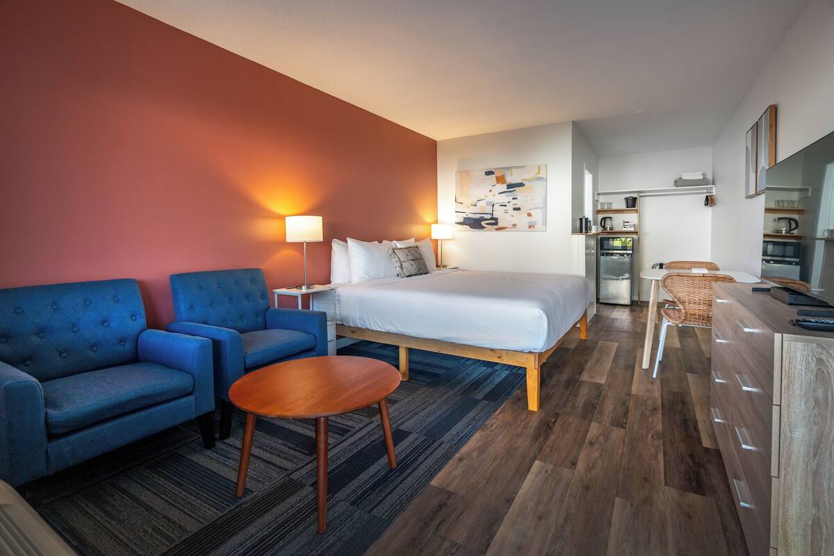 A hotel room with a bed, two blue sofas, a wooden coffee table, and a kitchenette in the background.