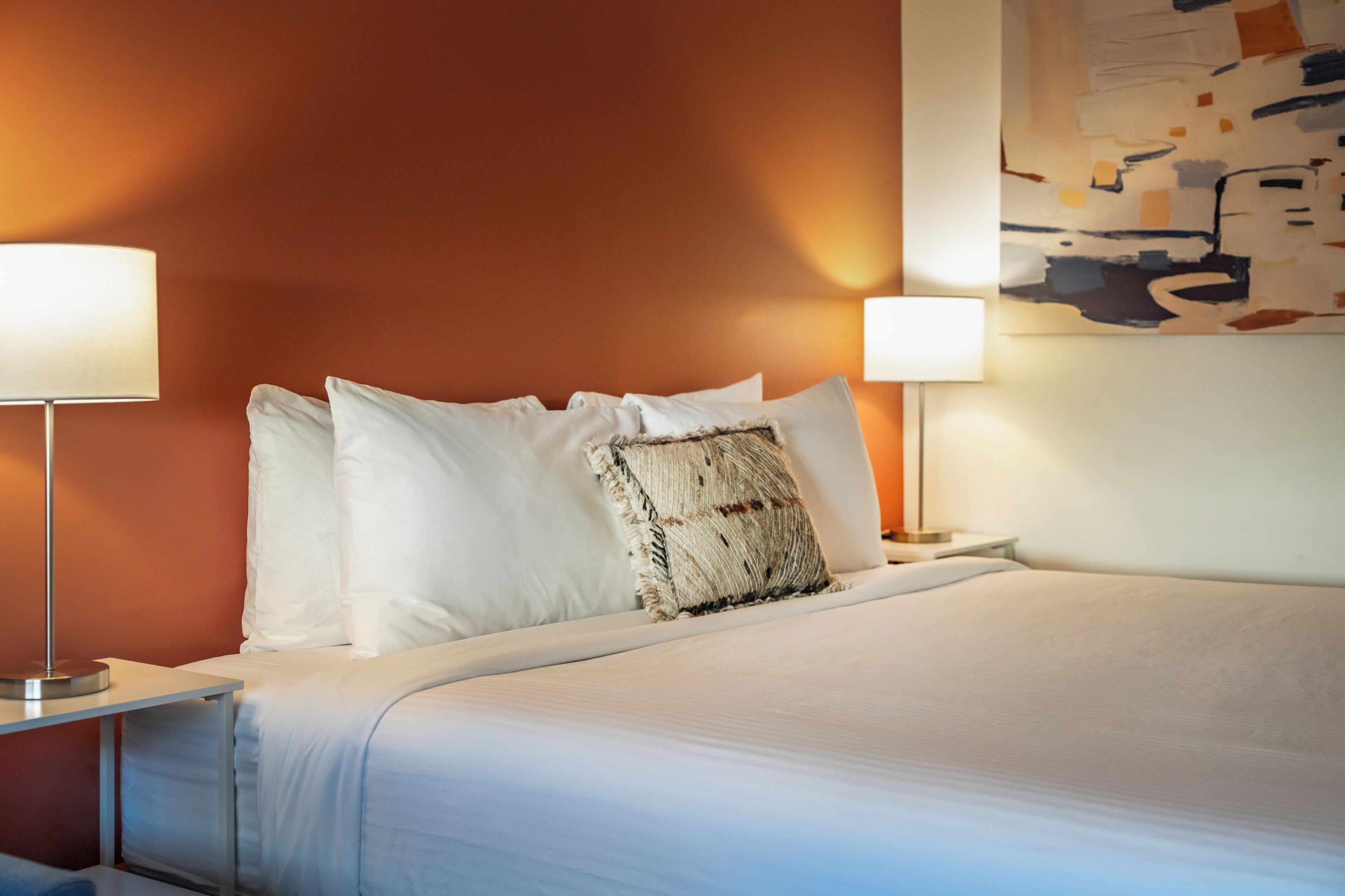 Een opgemaakt bed met witte lakens en een gestructureerd sierkussen, met aan weerszijden een nachtlampje op een nachtkastje.