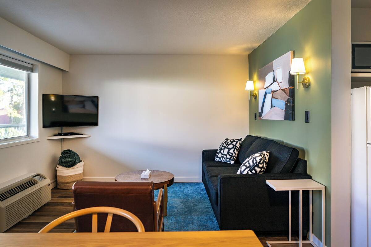 A compact living room with a flat-screen TV, a sofa with patterned cushions, a small table, and a blue carpet.