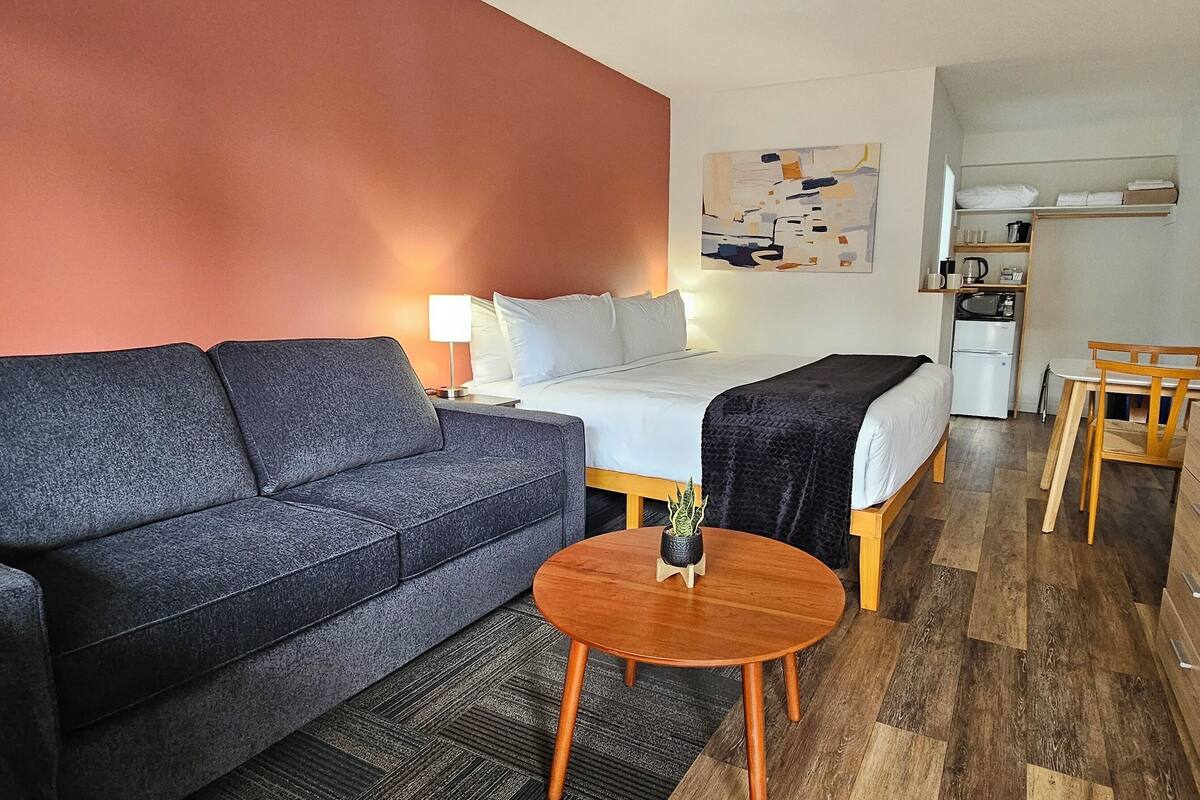 A modern hotel room with a grey sofa, a single bed, a wooden coffee table, a TV, and a kitchenette in the background.