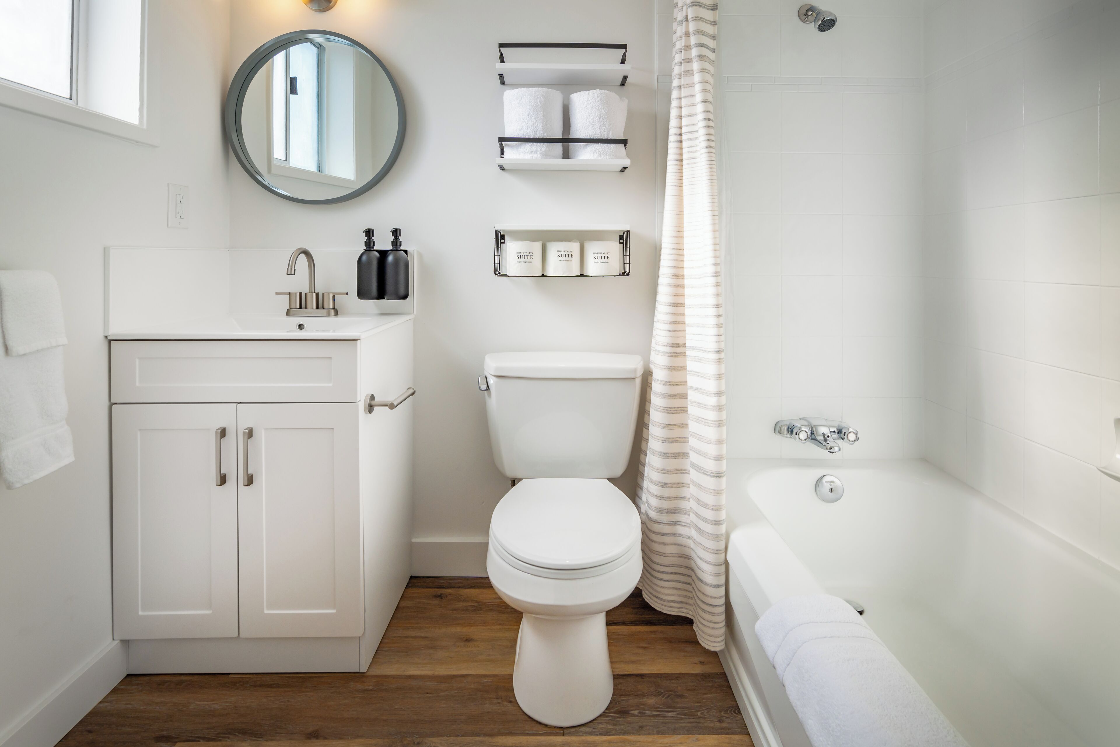Un baño moderno con un lavamanos, inodoro y bañera blancos. Hay repisas sobre el inodoro con artículos de aseo.