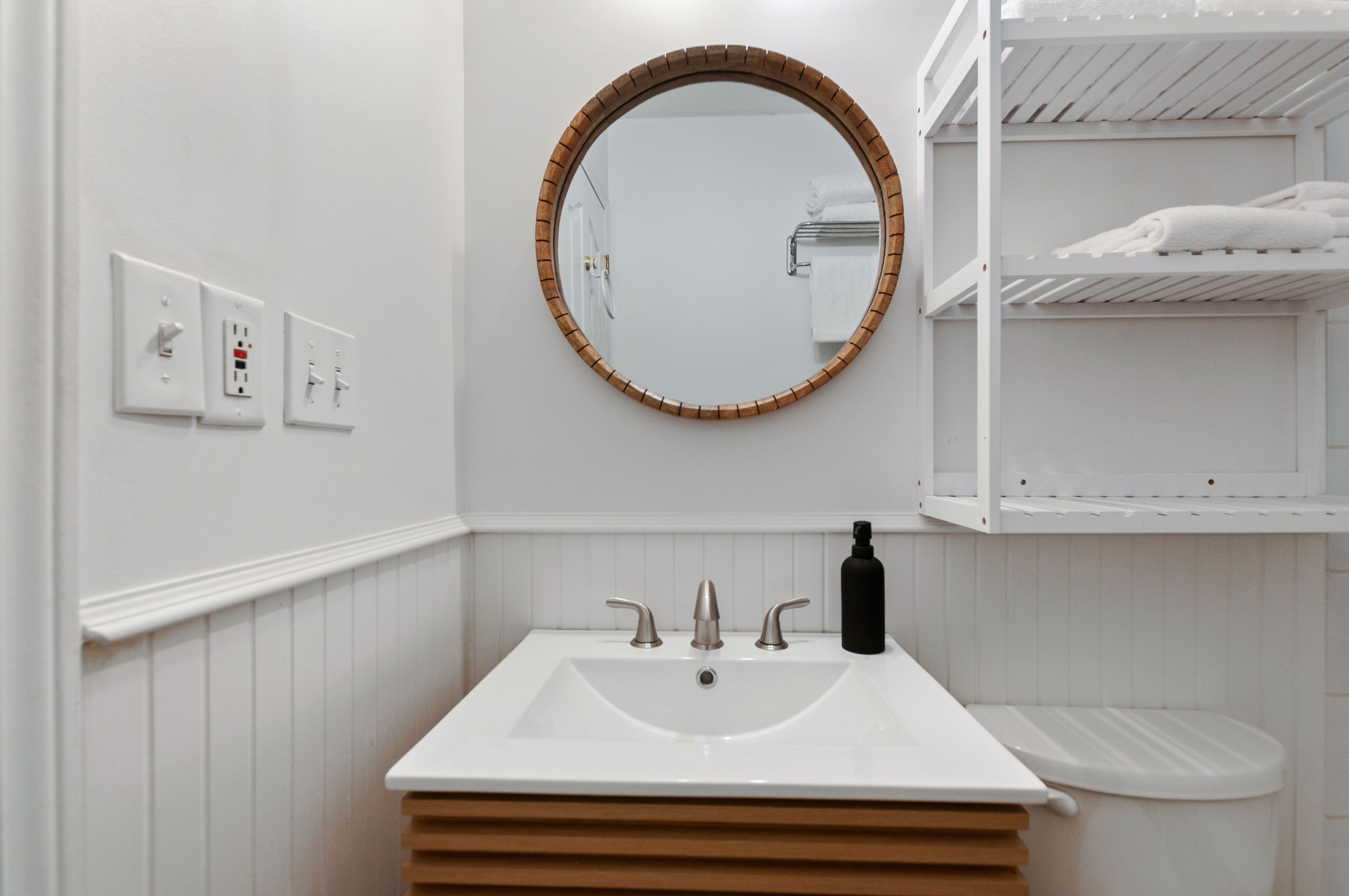 Un baño con un lavabo blanco, un espejo redondo con marco de madera y un inodoro blanco. Hay tres tomas de corriente en la pared y un estante con toallas sobre el inodoro.