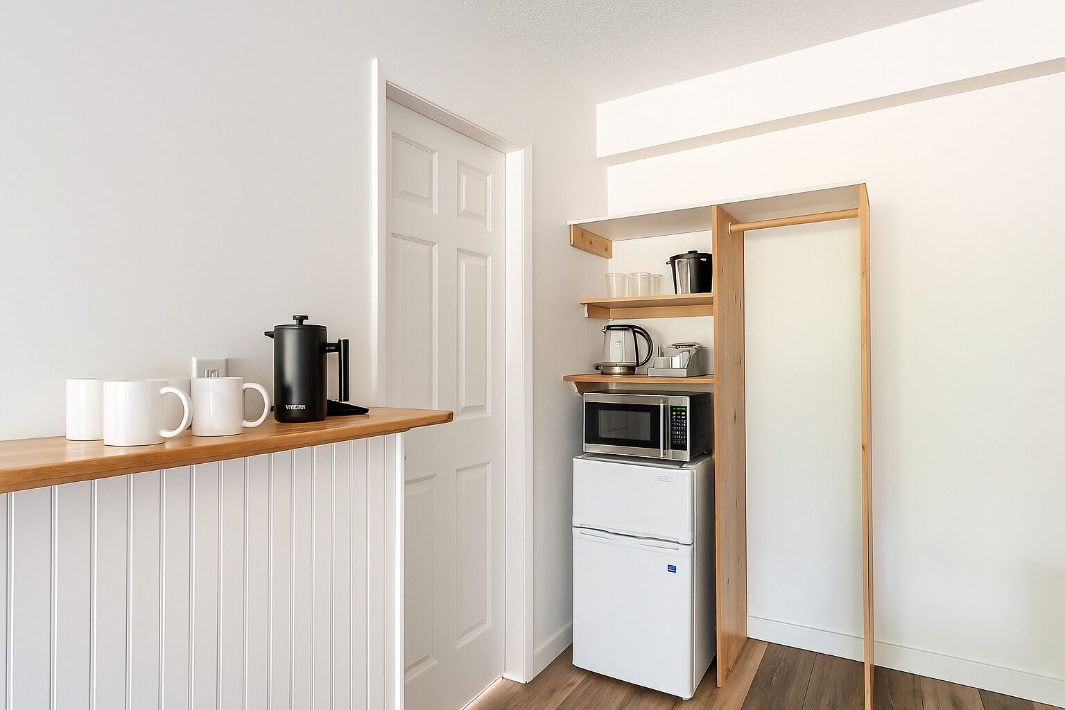 A compact kitchen with white cabinetry, a microwave, a coffee maker, and a kettle on a wooden shelf.