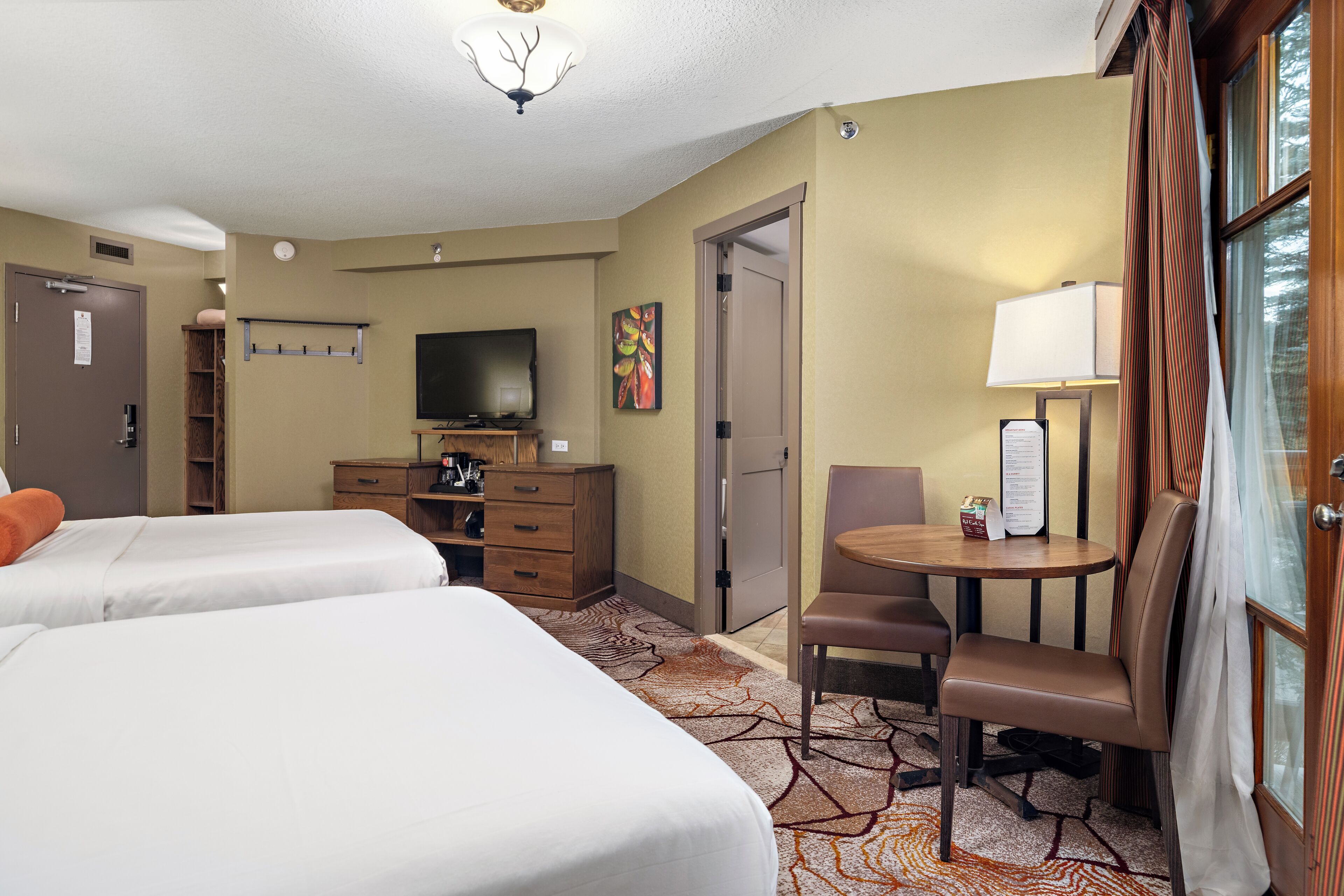 A hotel room with two beds, a television, a desk with a chair, a coffee machine, and a window with curtains.