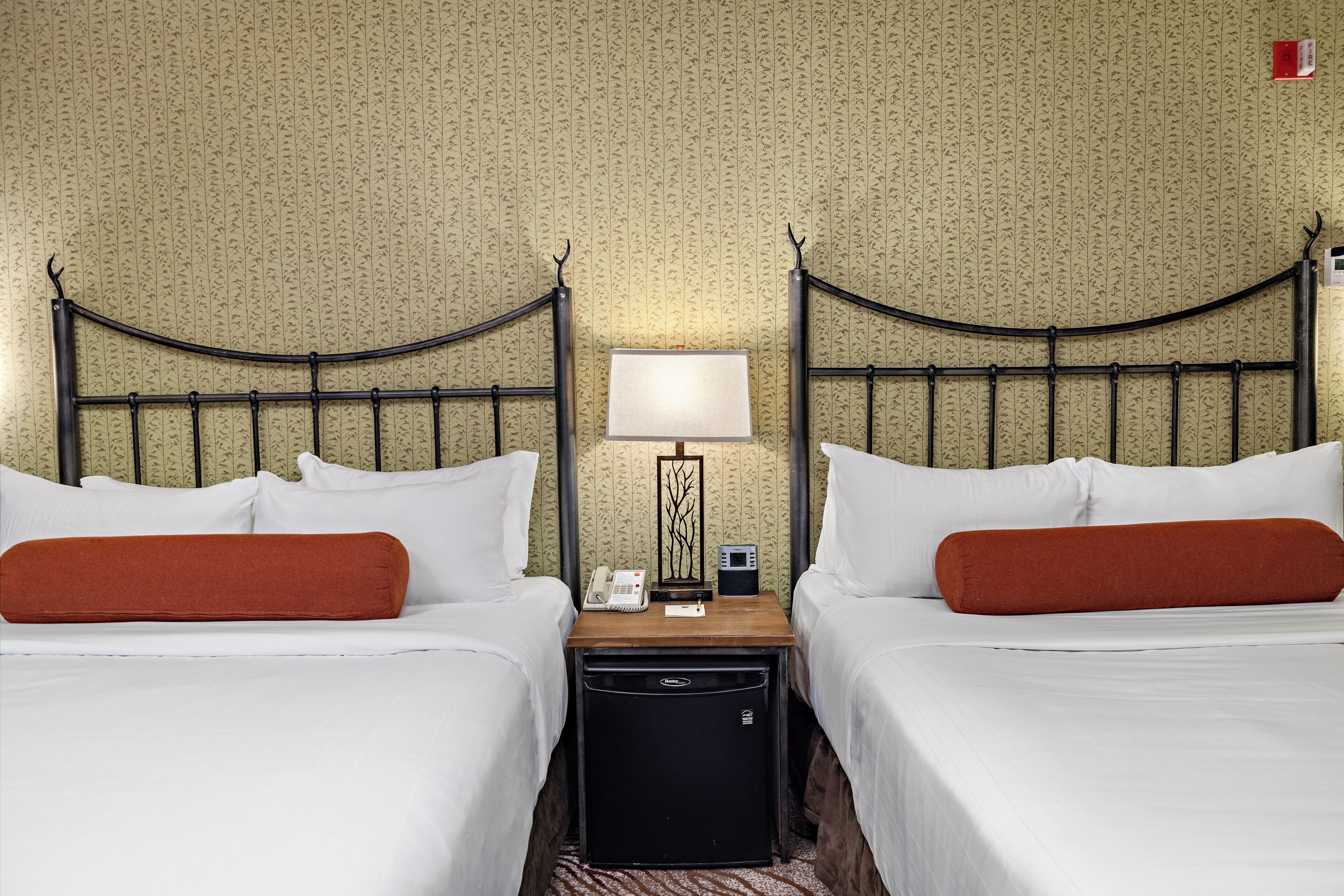 A hotel room with two beds, a nightstand with a lamp, a telephone, and a small black appliance.