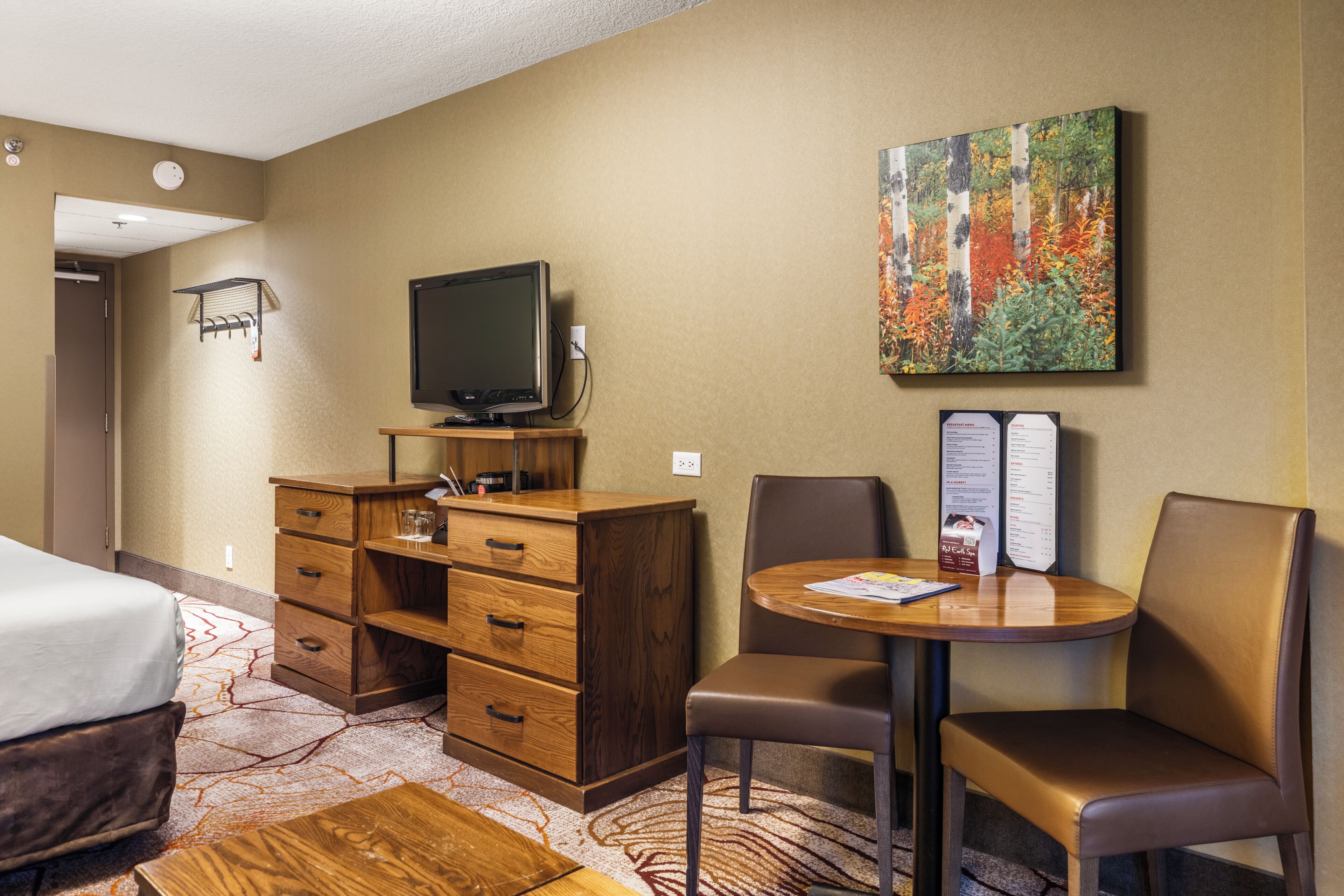 A hotel room with a bed, a wooden dresser, a television, a dining table with chairs, and a painting on the wall.