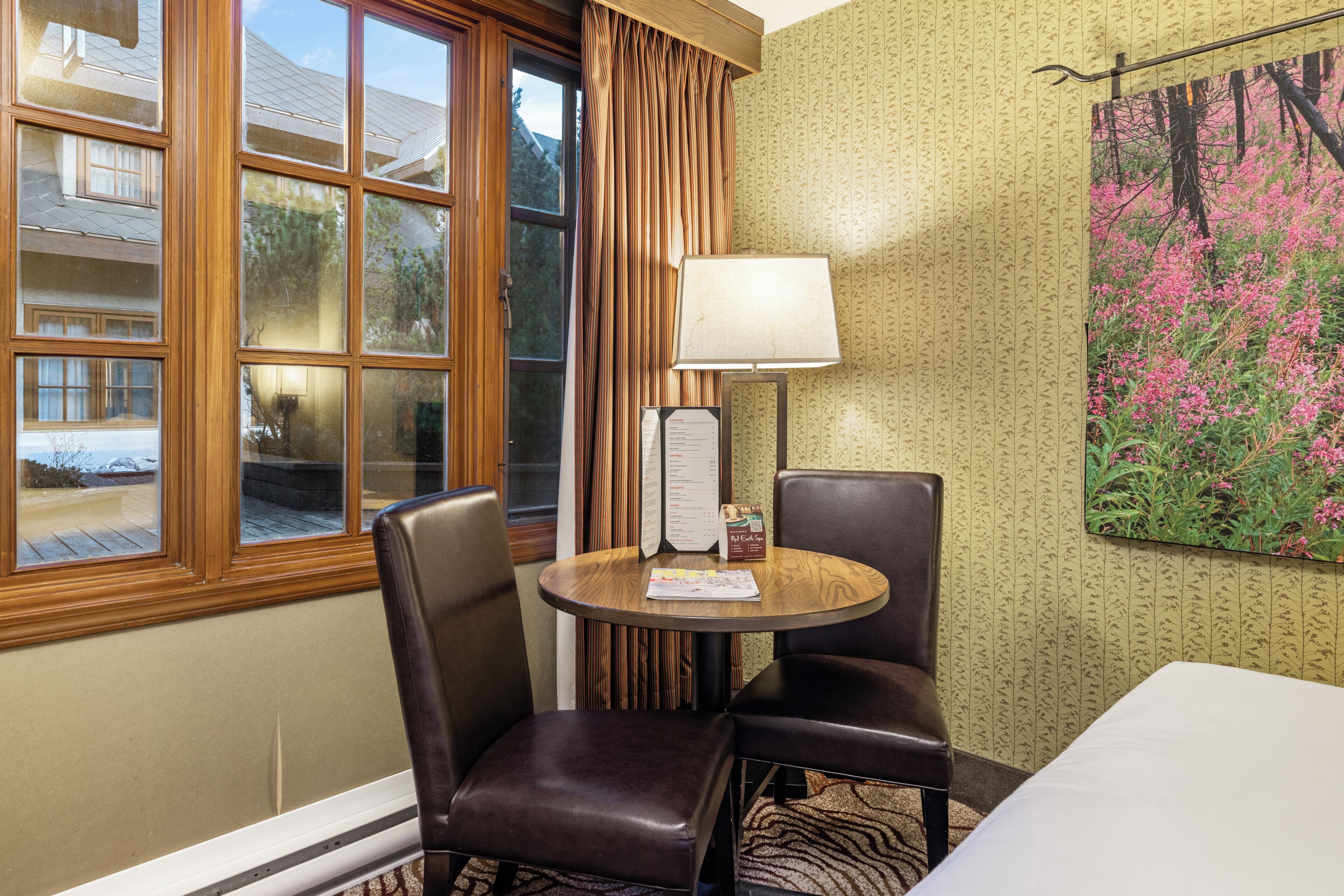 A hotel room with a large window, a table with a lamp, two chairs, and a menu stand.