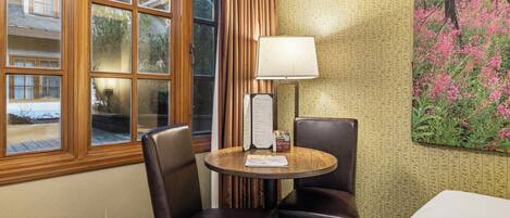 A hotel room with a large window, a table with a lamp, two chairs, and a menu stand.