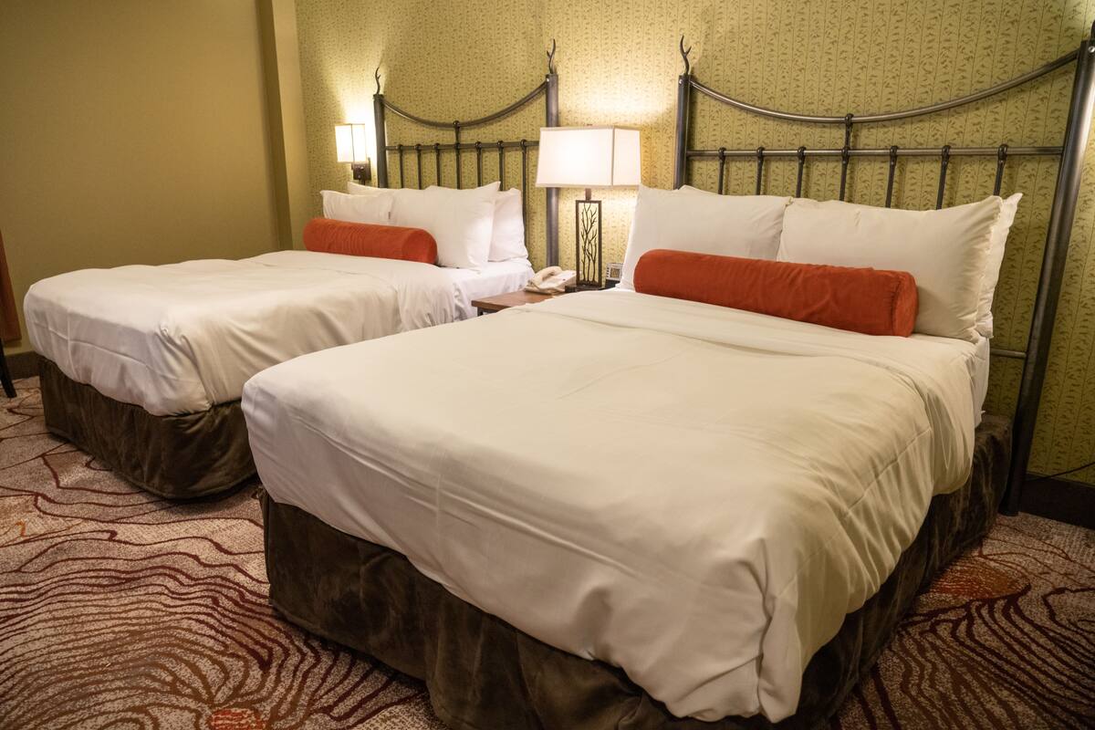 Habitación de hotel con dos camas, cada una con ropa de cama blanca y almohadas naranjas. Entre las camas, hay una mesita de noche con una lámpara y un teléfono. La habitación tiene una alfombra estampada y un cabecero de metal.
