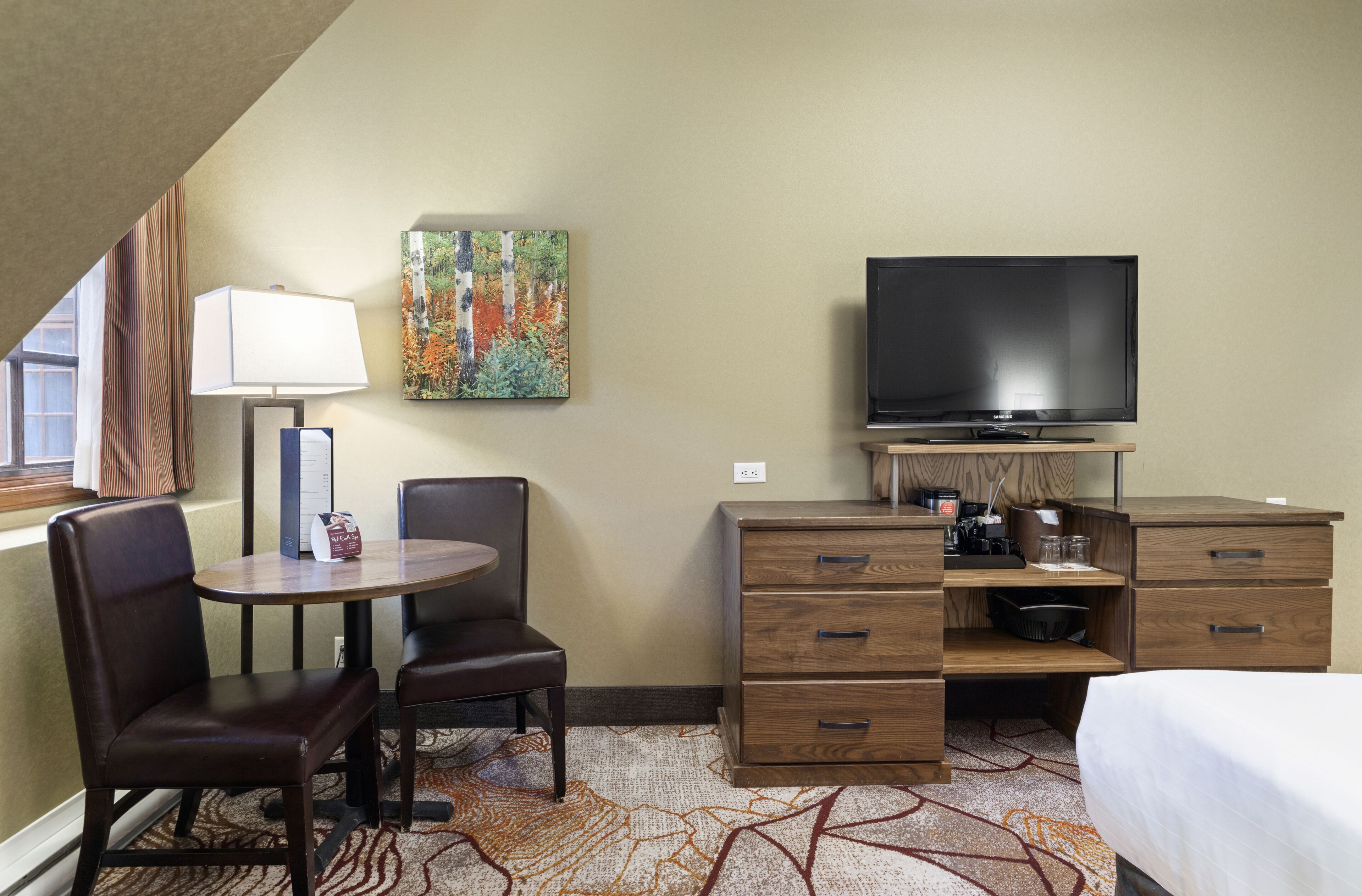 Une chambre d’hôtel avec un lit, un bureau et une chaise, un téléviseur, un tableau au mur et une fenêtre avec des rideaux.