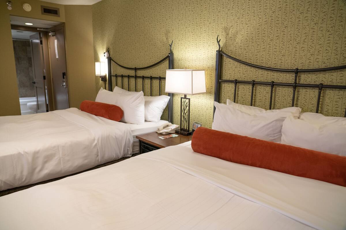 Habitación de hotel con dos camas, una mesita de noche, una lámpara y una puerta que da a un baño.