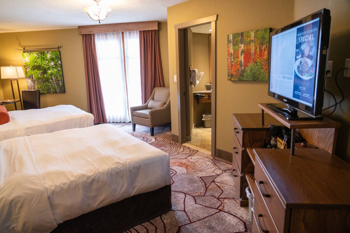 Habitación de hotel con dos camas, televisión, una silla y un baño visible a través de una puerta abierta.