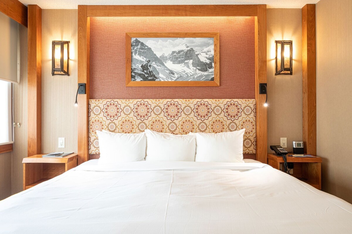 A hotel room with a large bed, a patterned headboard, two bedside tables with lamps, a telephone, and a framed picture on the wall.