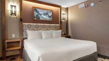 A hotel room with a large bed, a wooden headboard, a framed picture of a mountain, and a bedside table with a lamp.