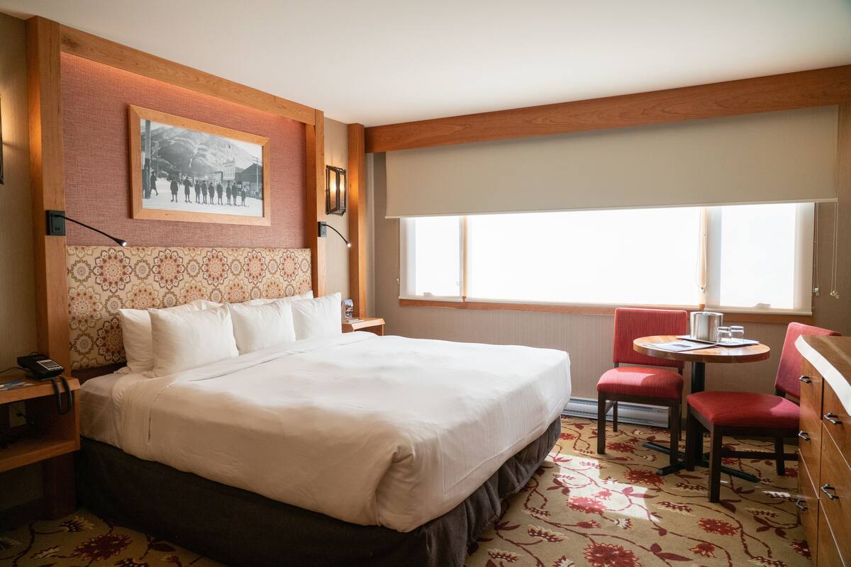 Habitación de hotel con una cama grande, una mesita con sillas, una ventana con vistas y un cuadro en la pared.