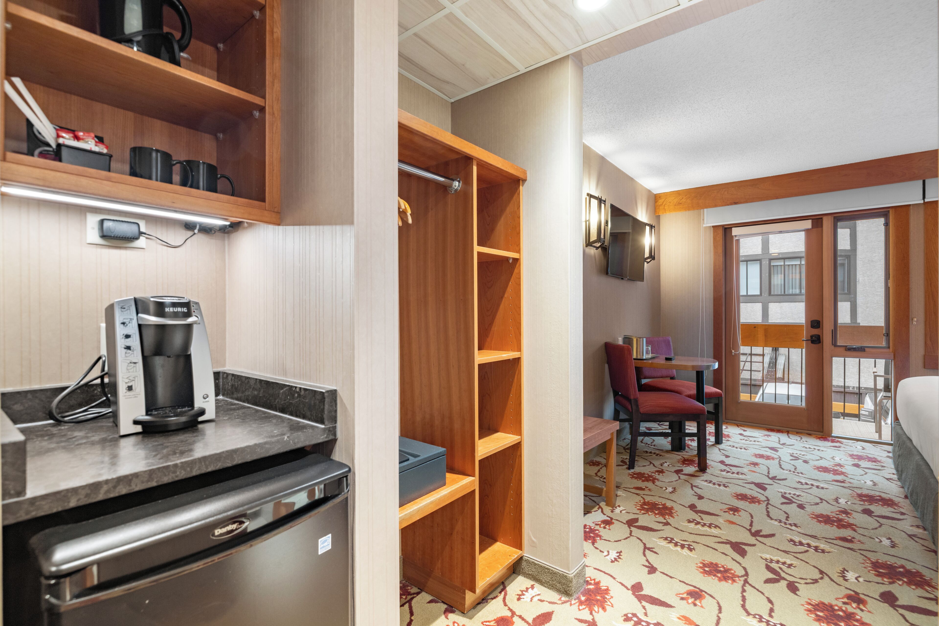 Une cuisine compacte équipée d’une machine à café, d’un réfrigérateur et d  ’un four à micro-ondes. On y trouve une étagère en bois et une petite table avec des chaises. La pièce est dotée d’un tapis à motifs et d’une porte donnant sur l’extérieur.