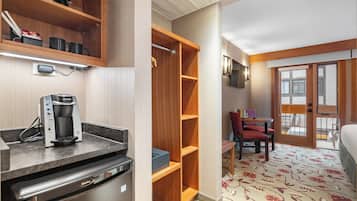 Cocina compacta con cafetera, refrigerador y microondas. Hay una estantería de madera y una pequeña mesa con sillas. La habitación tiene una alfombra estampada y una puerta que da al exterior.