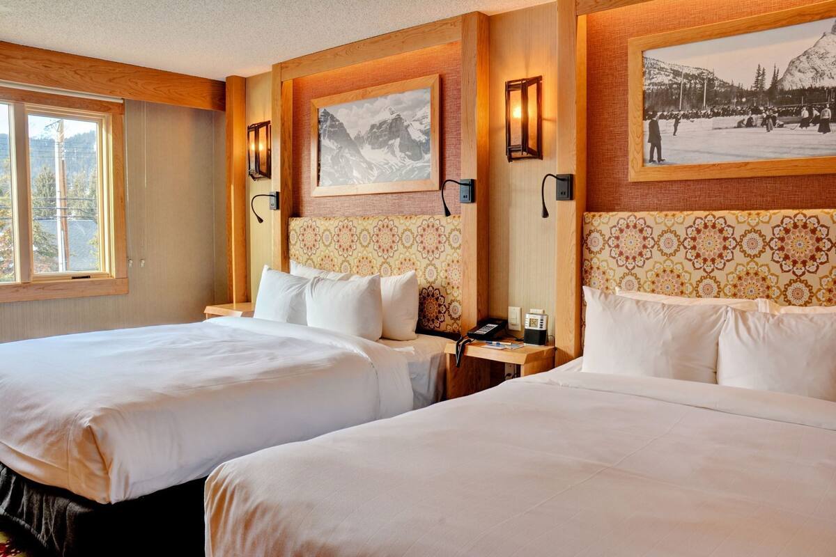 Habitación de hotel con dos camas, cabeceros de madera, lámparas fijadas a la pared, un cuadro enmarcado y una ventana.