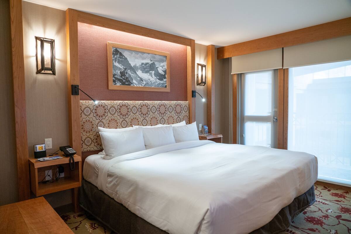 Habitación de hotel con una cama grande, cabecera de madera, un cuadro en la pared y ventana con vistas.